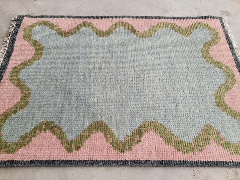 May include: A rectangular area rug featuring a central light blue field surrounded by a wavy olive green border and a pink outer frame. The rug is finished with a dark gray edge and short fringe on the left and right sides, showcasing a textured pile.