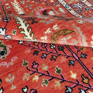 May include: A woven rug featuring a vibrant terracotta red background with a repeating pattern of stylized trees and floral motifs in white, navy blue, green, and pink. The rug is folded over, revealing a textured surface and a decorative border.