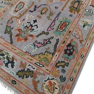 May include: A rectangular, gray area rug with an intricate, colorful pattern. The rug features a central field of gray with a repeating design of floral and geometric shapes in shades of orange, green, pink, and blue. A border frames the design.