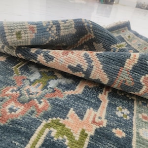 May include: A rolled-up, intricately patterned rug with a predominantly blue background. The design incorporates shades of pink, green, and cream, creating a detailed and textured appearance. The rug's edges are visible, showcasing its construction.