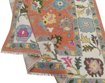 May include: A detailed rug with a folded corner, displaying a mix of colours. The rug has a terracotta base with floral patterns in pink, blue, yellow, and green, framed by a light blue and cream border. The rug's fringe is visible.