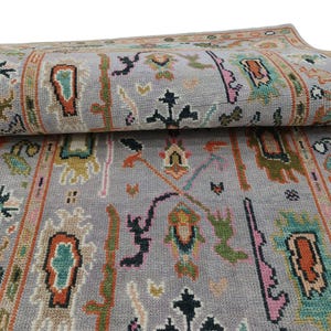 May include: A rolled-up, gray area rug with a detailed, colorful pattern. The design includes geometric shapes, floral motifs, and borders in shades of orange, green, pink, and black. The rug is made of a soft, woven material.