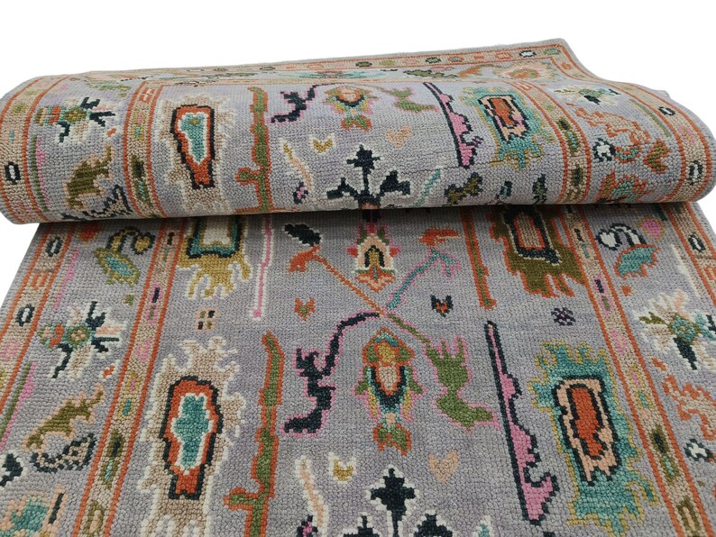May include: A rolled-up, gray area rug with a detailed, colorful pattern. The design includes geometric shapes, floral motifs, and borders in shades of orange, green, pink, and black. The rug is made of a soft, woven material.