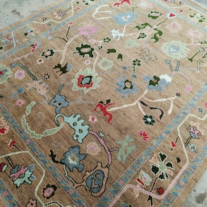 May include: A rectangular area rug featuring a tan background with a multi-colored floral pattern. The design includes stylized flowers and vines in shades of light blue, dark green, pink, and red, framed by a decorative border in blue and tan.