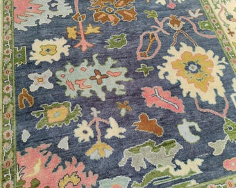 May include: A hand-knotted area rug featuring a deep blue background with an intricate pattern of stylized floral and organic shapes in shades of pink, yellow, light green, and cream. The rug includes a decorative border with green accents.
