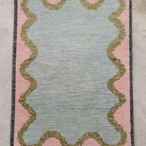 May include: A rectangular area rug featuring a central light blue field with a wavy, scalloped olive green border. The rug is framed by a solid pink background and a dark charcoal gray outer edge, finished with short white fringe on both ends.