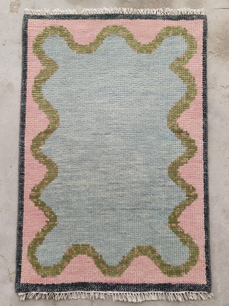 May include: A rectangular area rug featuring a central light blue field with a wavy, scalloped olive green border. The rug is framed by a solid pink background and a dark charcoal gray outer edge, finished with short white fringe on both ends.