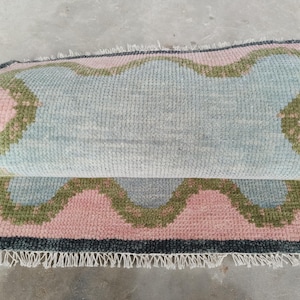 May include: A rectangular area rug featuring a light blue center with a wavy, scalloped border in olive green and soft pink. The rug has a dark gray outer edge and white fringe along the bottom. The rug is folded over to show the textured pile.