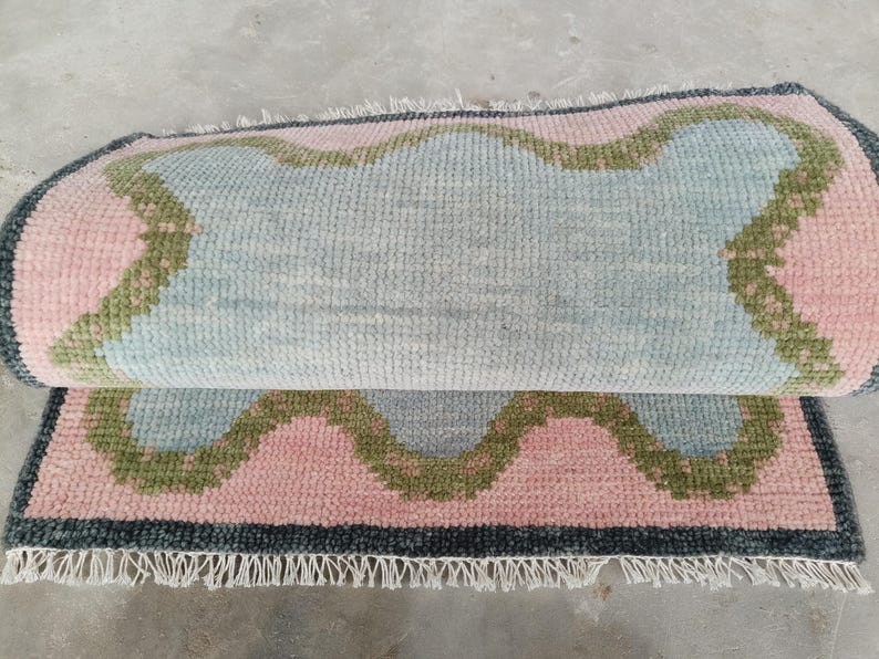May include: A rectangular area rug featuring a light blue center with a wavy, scalloped border in olive green and soft pink. The rug has a dark gray outer edge and white fringe along the bottom. The rug is folded over to show the textured pile.