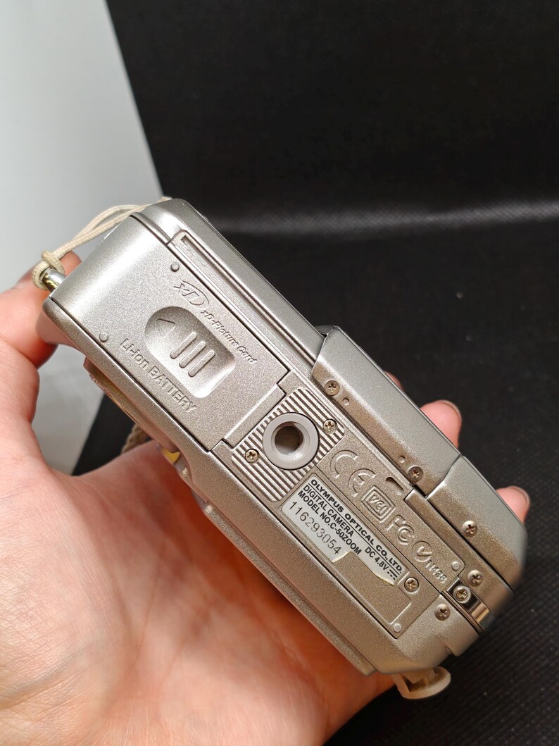 以下が含まれることがあります： シルバーのオリンパスC-50ズームデジタルカメラ。カメラはシルバーのボディで、「OLYMPUS OPTICAL CO., LTD. DIGITAL CAMERA MODEL NO. C-50 ZOOM DC 4.8V」と書かれたラベルが付いています。カメラは手で持たれています。