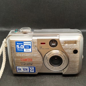 以下が含まれることがあります： シルバーのオリンパスCamedia C-50 Zoomデジタルカメラ。5.0メガピクセル、3倍光学ズーム、充電式リチウムイオンバッテリーを搭載。カメラはシルバーのボディで、レンズとリストストラップが付いています。