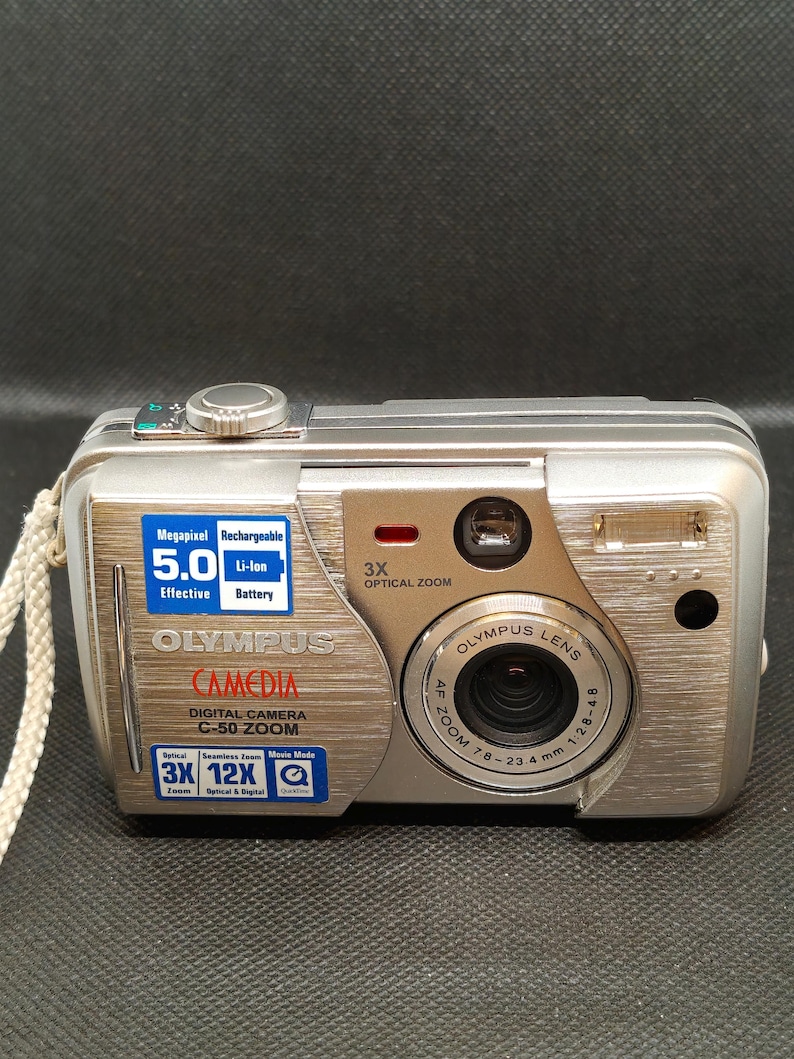 以下が含まれることがあります： シルバーのオリンパスCamedia C-50 Zoomデジタルカメラ。5.0メガピクセル、3倍光学ズーム、充電式リチウムイオンバッテリーを搭載。カメラはシルバーのボディで、レンズとリストストラップが付いています。