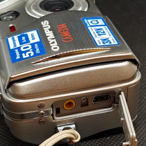 以下が含まれることがあります： シルバーのオリンパスデジタルカメラ。有効5.0メガピクセルのバッテリーを搭載。ズームレンズとストラップが付いています。カメラのポートカバーが開いており、ポートが見えています。