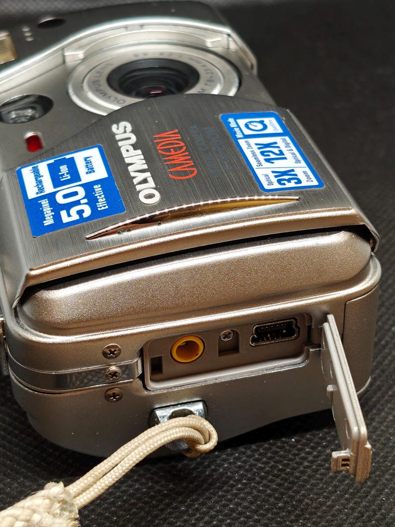 以下が含まれることがあります： シルバーのオリンパスデジタルカメラ。有効5.0メガピクセルのバッテリーを搭載。ズームレンズとストラップが付いています。カメラのポートカバーが開いており、ポートが見えています。