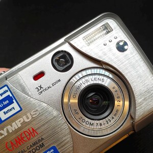 以下が含まれることがあります： シルバーのオリンパスCamedia C-50 Zoomデジタルカメラ。3倍光学ズーム、AFズームレンズ、内蔵フラッシュなどの機能を備えています。カメラには、「Li-Ion Battery」や「QuickTime」などのテキストが記載されたステッカーがいくつかあります。