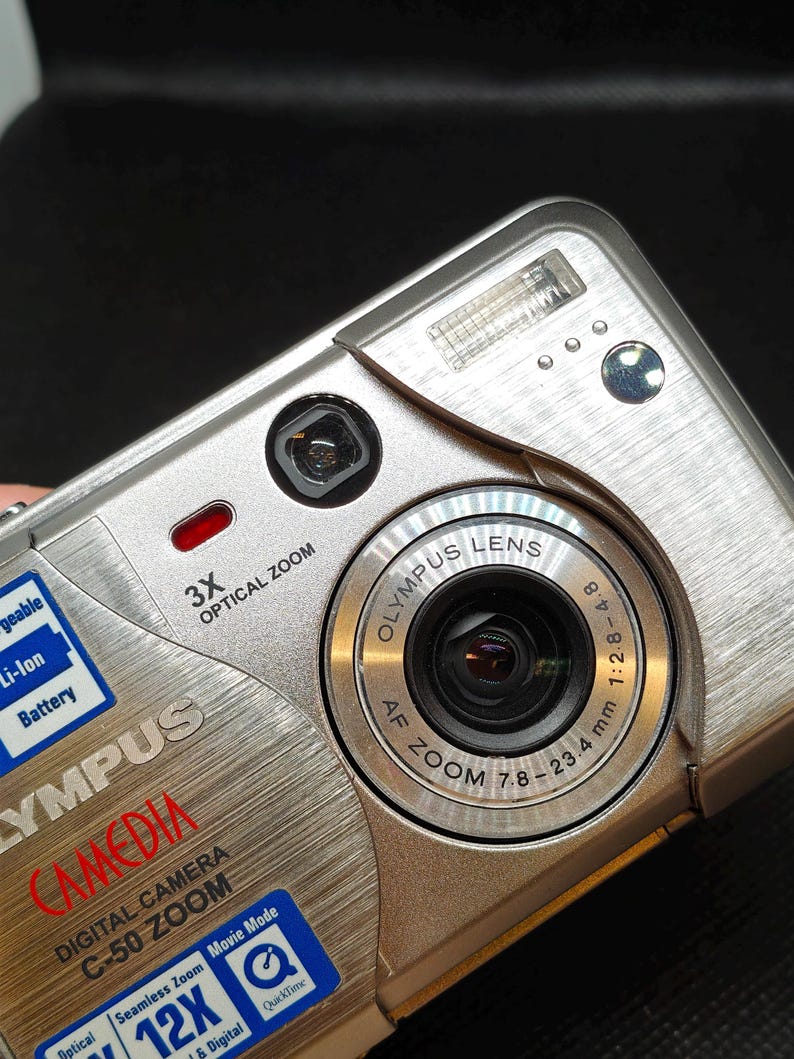 以下が含まれることがあります： シルバーのオリンパスCamedia C-50 Zoomデジタルカメラ。3倍光学ズーム、AFズームレンズ、内蔵フラッシュなどの機能を備えています。カメラには、「Li-Ion Battery」や「QuickTime」などのテキストが記載されたステッカーがいくつかあります。