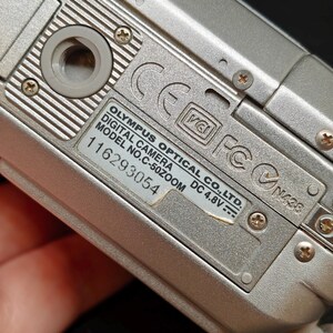 以下が含まれることがあります： 銀色のオリンパスデジタルカメラのクローズアップ。カメラの底面が見え、モデル番号C-50ZOOMやその他の技術的な詳細が表示されています。カメラは金属仕上げで、複数のネジで固定されています。