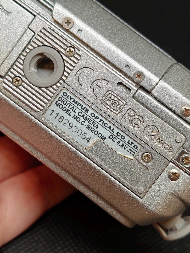 以下が含まれることがあります： 銀色のオリンパスデジタルカメラのクローズアップ。カメラの底面が見え、モデル番号C-50ZOOMやその他の技術的な詳細が表示されています。カメラは金属仕上げで、複数のネジで固定されています。