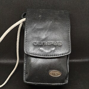 以下が含まれることがあります： 前面に「OLYMPUS」のブランド名がエンボス加工された黒革のカメラケース。ケースはフラップ開閉式で、「CAMEDIA」と書かれた小さな金属製のエンブレムが付いています。白いコードが側面に付いています。