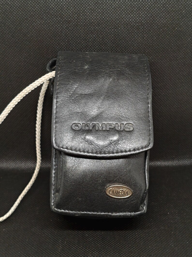 以下が含まれることがあります： 前面に「OLYMPUS」のブランド名がエンボス加工された黒革のカメラケース。ケースはフラップ開閉式で、「CAMEDIA」と書かれた小さな金属製のエンブレムが付いています。白いコードが側面に付いています。