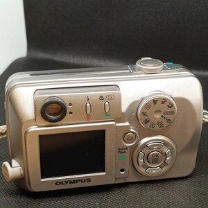 以下が含まれることがあります： 小さなLCD画面とさまざまなボタンとダイヤルを備えた、シルバーのオリンパスデジタルカメラ。カメラにはレンズ、フラッシュがあり、前面には「OLYMPUS」の文字が印刷されています。ベージュのストラップが側面に付いています。