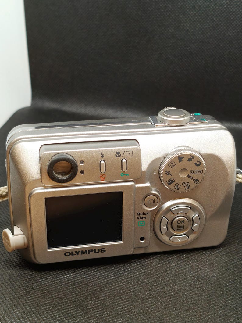 以下が含まれることがあります： 小さなLCD画面とさまざまなボタンとダイヤルを備えた、シルバーのオリンパスデジタルカメラ。カメラにはレンズ、フラッシュがあり、前面には「OLYMPUS」の文字が印刷されています。ベージュのストラップが側面に付いています。
