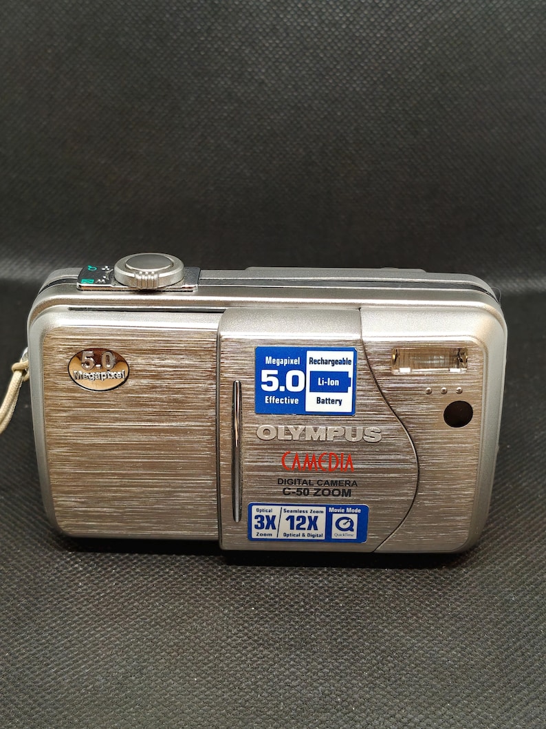 以下が含まれることがあります： シルバーのオリンパスC-50 ZOOMデジタルカメラ。5.0メガピクセルの解像度と充電式バッテリーを搭載。3倍光学ズームと12倍デジタルズームを搭載。ヘアライン仕上げの金属製です。