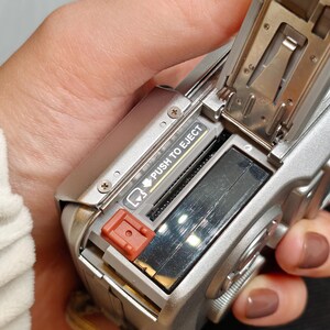 以下が含まれることがあります： バッテリーコンパートメントが開いた、シルバーのデジタルカメラ。バッテリーのスロットと赤いイジェクトボタンが見えます。「PUSH TO EJECT」の文字が見えます。