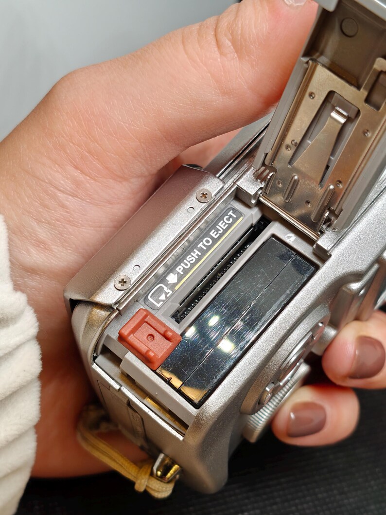 以下が含まれることがあります： バッテリーコンパートメントが開いた、シルバーのデジタルカメラ。バッテリーのスロットと赤いイジェクトボタンが見えます。「PUSH TO EJECT」の文字が見えます。