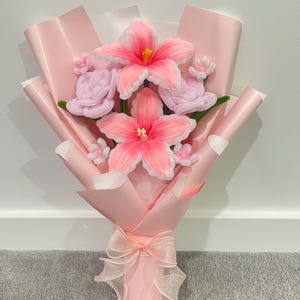 May include: A handmade bouquet of pink yarn flowers, including large hibiscus-like blooms and smaller rose-like buds. The flowers are wrapped in pink paper and tied with a sheer ribbon.