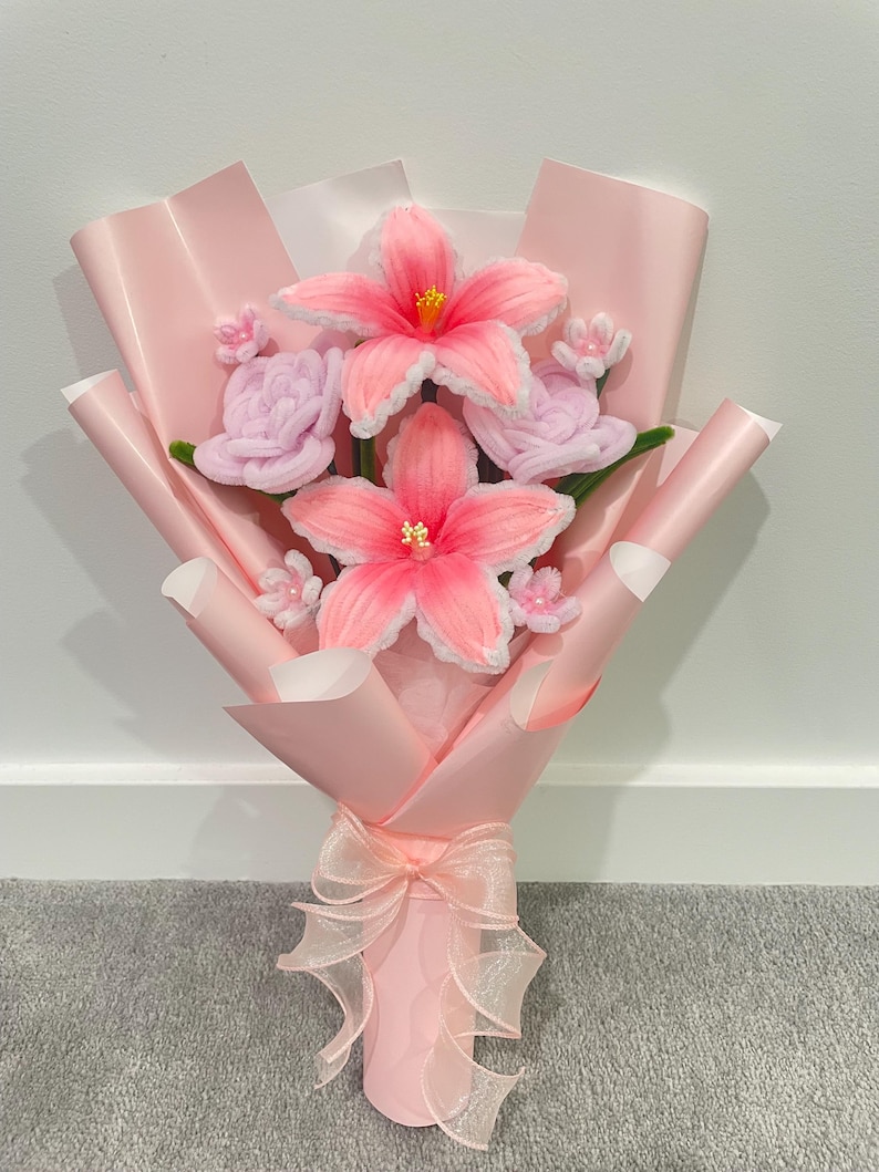 May include: A handmade bouquet of pink yarn flowers, including large hibiscus-like blooms and smaller rose-like buds. The flowers are wrapped in pink paper and tied with a sheer ribbon.