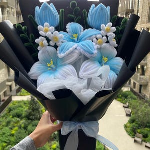 May include: A bouquet of handcrafted pipe cleaner flowers featuring light blue tulips, white and blue lilies, and small white blossoms with yellow centres. The arrangement is wrapped in black paper with a light blue ribbon tied at the base.