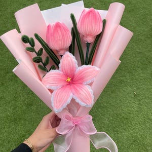 May include: A handmade bouquet of pink yarn flowers, including tulips and a large flower with white edging. The bouquet is wrapped in pink paper and tied with a sheer pink ribbon. Green stems and leaves provide contrast.