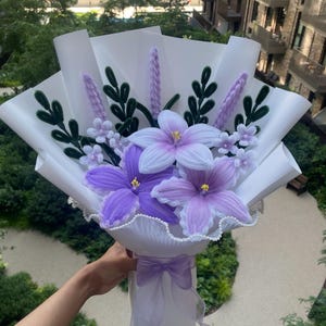 May include: A bouquet of handcrafted pipe cleaner flowers featuring purple and white lilies, small light purple blossoms, and dark green leafy stems. The arrangement is wrapped in white paper with a sheer light purple ribbon and the text Bouquetsbytasnim.