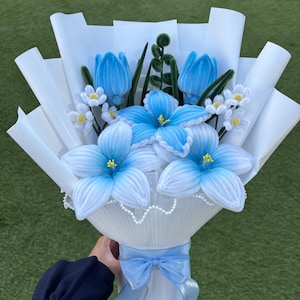 May include: A bouquet of handcrafted blue and white flowers made from pipe cleaners. The arrangement includes blue tulips, lilies, and small white blossoms with yellow centres, wrapped in white paper with a light blue ribbon tied at the base.