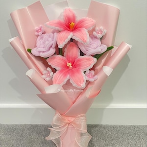 May include: A bouquet of pipe cleaner flowers featuring two large pink lilies with yellow centres, two light purple roses, and small white blossoms. The arrangement is wrapped in light pink paper and tied with a sheer ribbon, with the text BouquetsByTasnim visible.
