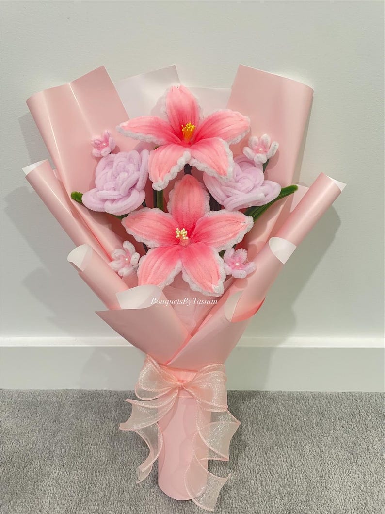 May include: A bouquet of pipe cleaner flowers featuring two large pink lilies with yellow centres, two light purple roses, and small white blossoms. The arrangement is wrapped in light pink paper and tied with a sheer ribbon, with the text BouquetsByTasnim visible.