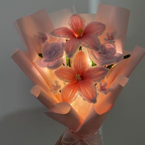 May include: A bouquet of crocheted pink lily and rose flowers wrapped in light pink paper. The arrangement features integrated warm white LED string lights that illuminate the petals from within, tied at the base with a sheer pink ribbon.