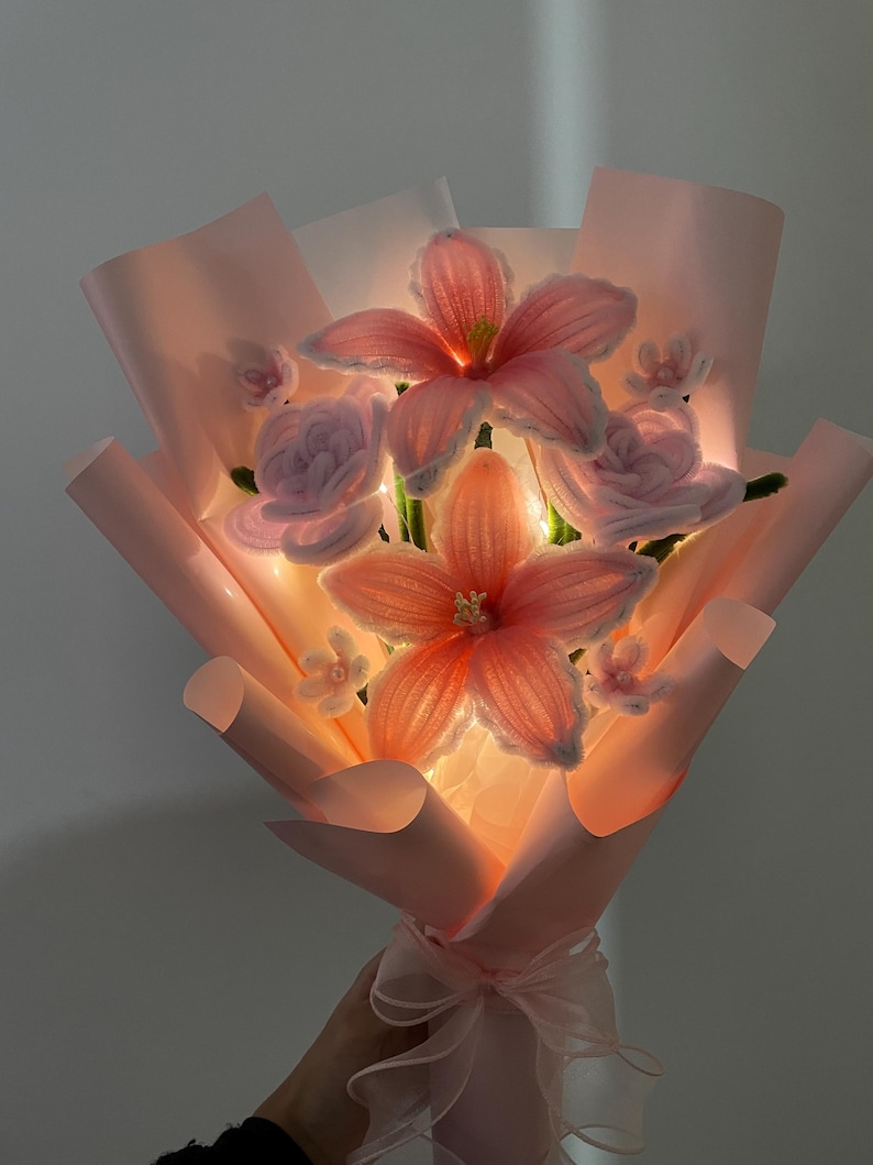 May include: A bouquet of crocheted pink lily and rose flowers wrapped in light pink paper. The arrangement features integrated warm white LED string lights that illuminate the petals from within, tied at the base with a sheer pink ribbon.