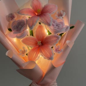 Op de afbeelding: Een boeket handgemaakte roze en perzik kleurige bloemen, verpakt in roze papier en vastgebonden met een doorschijnend lint. De bloemen hebben een delicate, gloeiende uitstraling, met zichtbare details in de bloemblaadjes en centra. Een uniek bloemstuk.