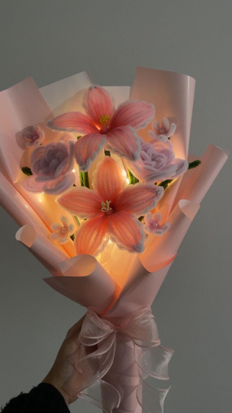 May include: A bouquet of handmade pink and peach-coloured flowers, wrapped in pink paper and tied with a sheer ribbon. The flowers have a delicate, glowing appearance, with visible details in the petals and centres. The bouquet is a unique floral arrangement.