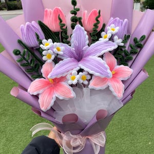 May include: A bouquet of handmade fabric flowers in shades of pink, purple, and white. The bouquet includes tulips, lilies, and small daisy-like flowers, wrapped in purple paper and tied with a pink ribbon. The flowers are arranged with green stems.