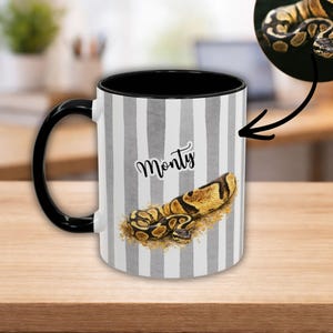 May include: A white ceramic mug with a black handle and interior. The mug features a vertical gray and white striped pattern. A detailed illustration of a snake is printed on the mug, along with the name "Monty" in a script font.