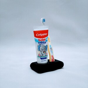 May include: A black rectangular holder with a tube of Colgate Baby Training toothpaste and a yellow toothbrush. The toothpaste tube is white with red and blue accents, featuring an elephant illustration. The toothbrush has a blue and white bristle head.