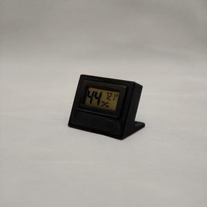 Puede incluir: Un termómetro e higrómetro digital negro muestra 44% y 72.1°F en una pequeña pantalla LCD. El dispositivo está alojado en un soporte negro en ángulo, diseñado para colocarse en posición vertical. El fondo es una tela lisa de color blanquecino. La temperatura es equivalente a 22.3°C.