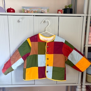 May include: A patchwork jacket in a variety of colours, including red, green, blue, yellow, and brown. The jacket has a round collar and button closure. It is hanging on a white hanger. The jacket is made of squares.