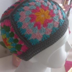 May include: A crochet beanie constructed from multiple square motifs featuring a vibrant granny square pattern in shades of yellow, pink, and light blue, all bordered by a solid dark grey yarn that forms the base and structural seams of the hat.