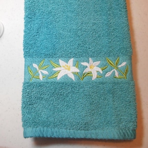 May include: A teal hand towel with a white embroidered border of lilies and green leaves.