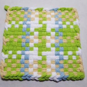 May include: A square, woven potholder in a checkerboard pattern. The potholder features a mix of green, white, light blue, and tan squares. The edges are finished with a braided border in green and tan. A small loop is attached to one corner.