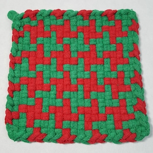May include: A square, woven potholder in a red and green pattern. The potholder is made of thick, woven threads, with a braided green border and a small loop for hanging. The pattern is a repeating design of red and green squares.
